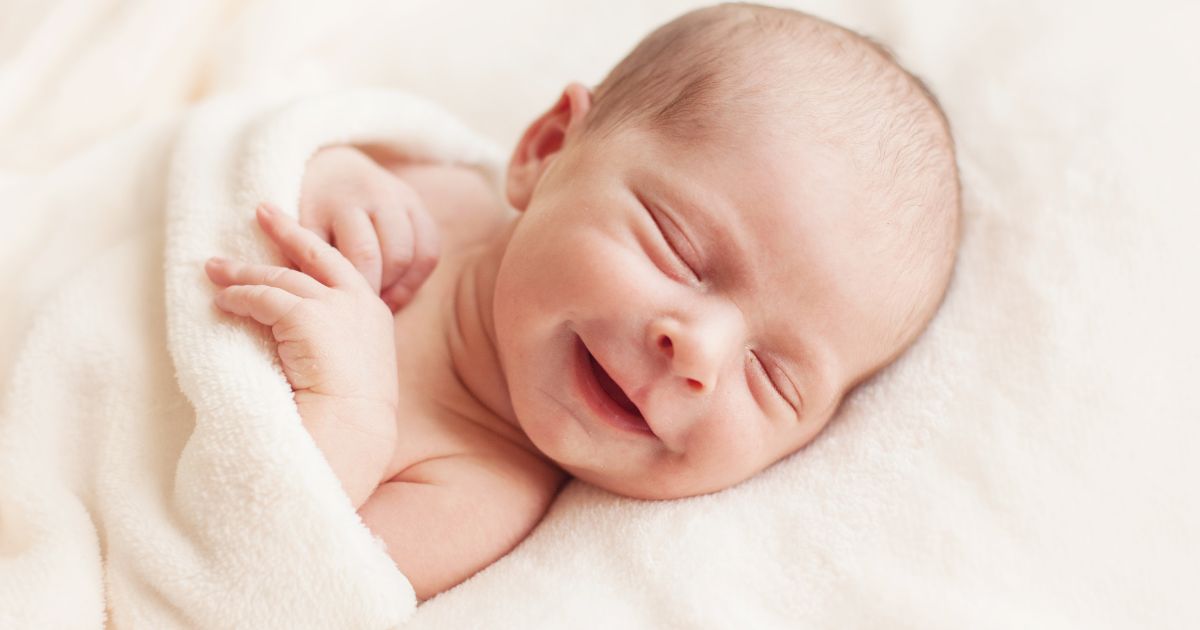 A newborn baby smiling while asleep, wrapped up in a cream coloured blanket